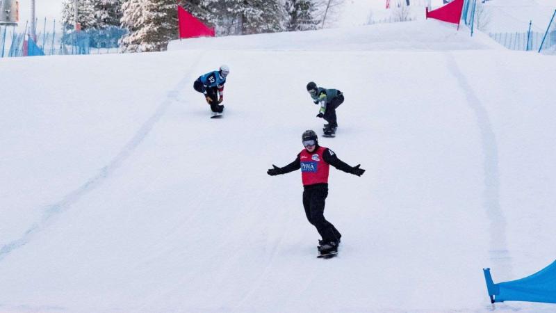 A male Para snowboarder crossing the finish line ahead of other two athletes