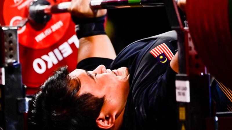 A man with an uniform with the flag of Malaysian pushing the bar on a bench press