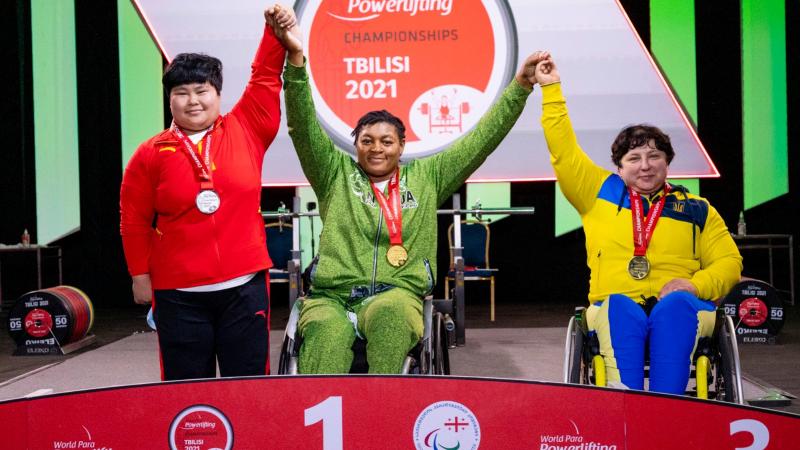 A woman in a wheelchair holding arms with a standing woman and another woman in a wheelchair in a podium