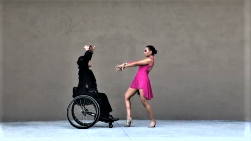 A male wheelchair dancer dressed in black dancing together with a standing woman in a pink dress