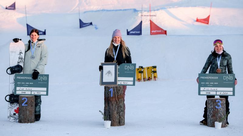 Three women smiling and showing checks in a snowboard medal ceremony in the snow