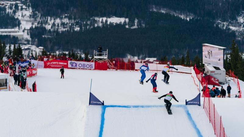 Four male athletes competing in a Para snowboard cross race. Three of them are jumping in the air, while the last one is about to jump.