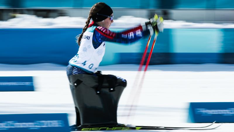 A woman in a sit ski competition pushing herself forward with two sticks.