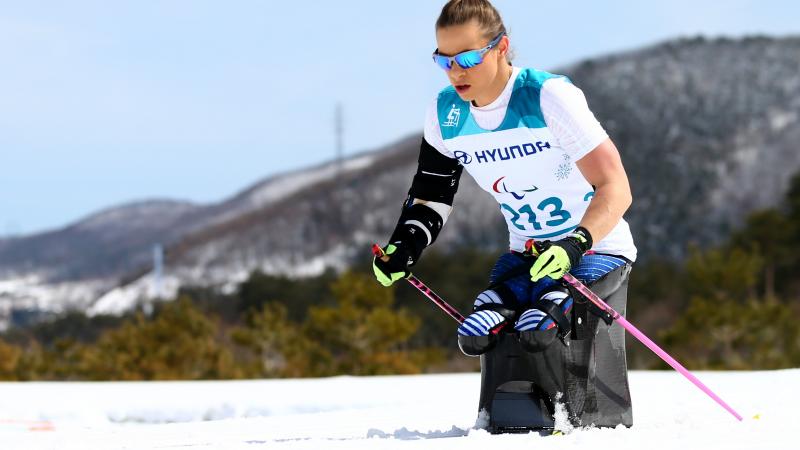 A woman with goggles pushing herself forward with two sticks in a sit ski competition on a sunny day.