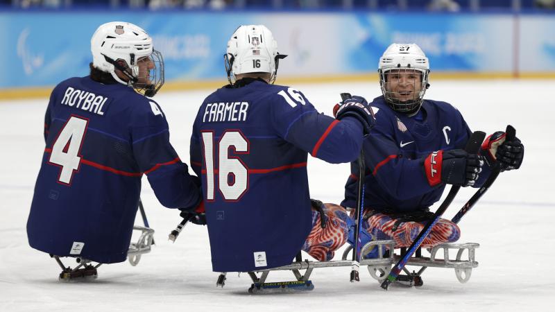 Three players in full Para ice hockey gear celebrating a goal with a smile.