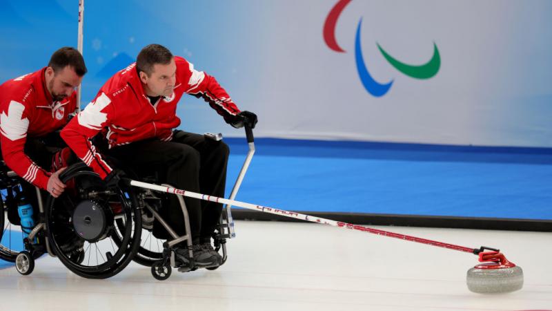 Mark ideson of Canada is held by a teammate as he goes to release the curling stone at Beijing 2022