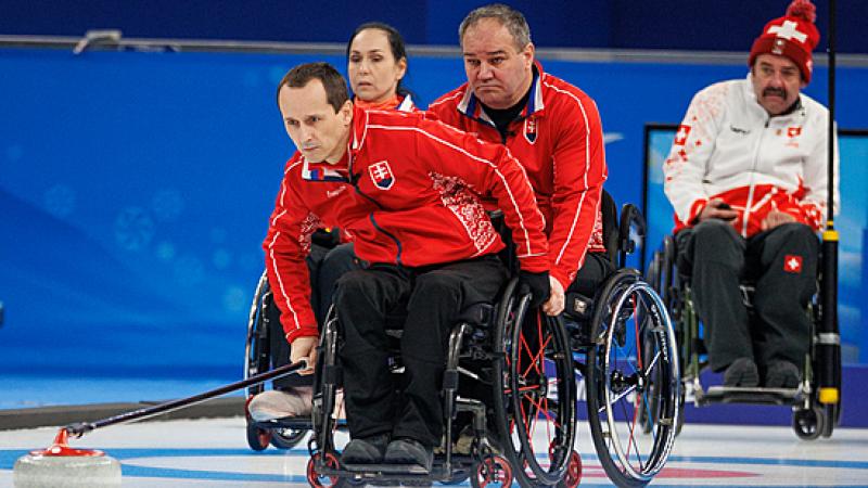 A Slovakian wheelchair curler pushes the stone whilst his wheelchair is held by a teammate