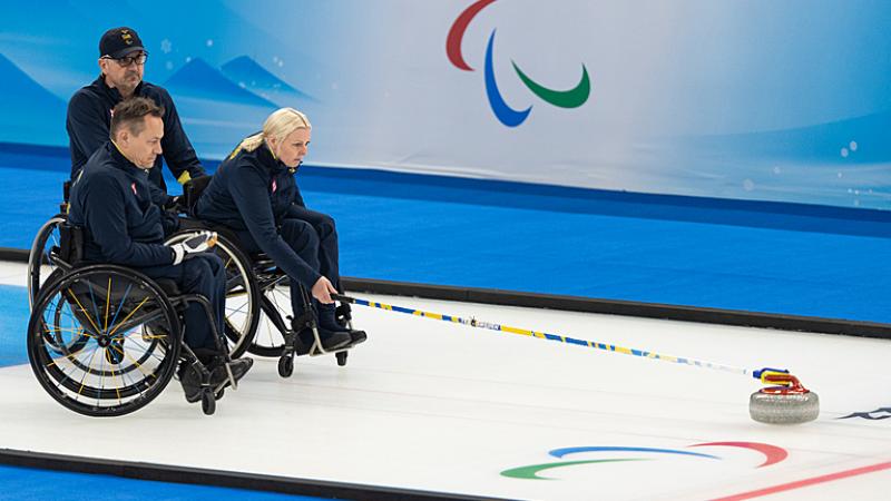 A Swedish wheelchair curler pushes the stone forward while her teammates looks on