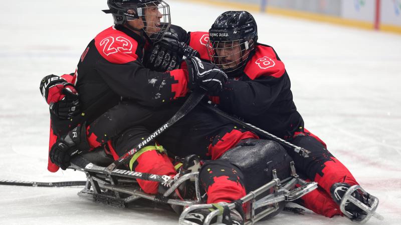Three men on sleds lying on the ice and hugging each other in full Para ice hockey gear.