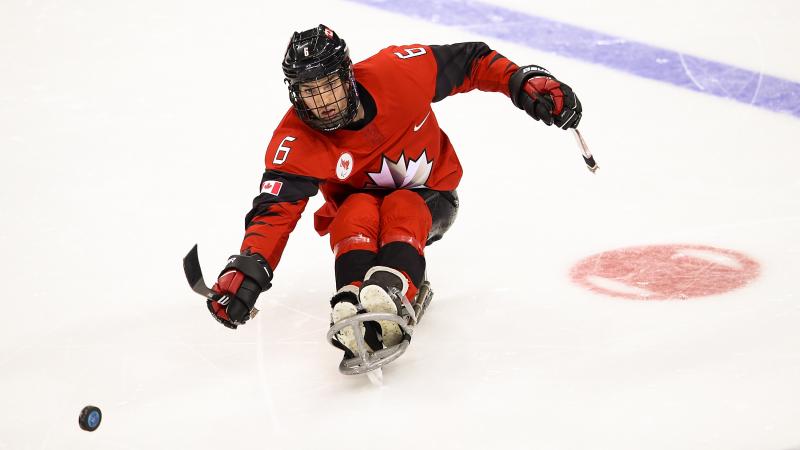 A male player on a sledge kicks the puck with a stick.