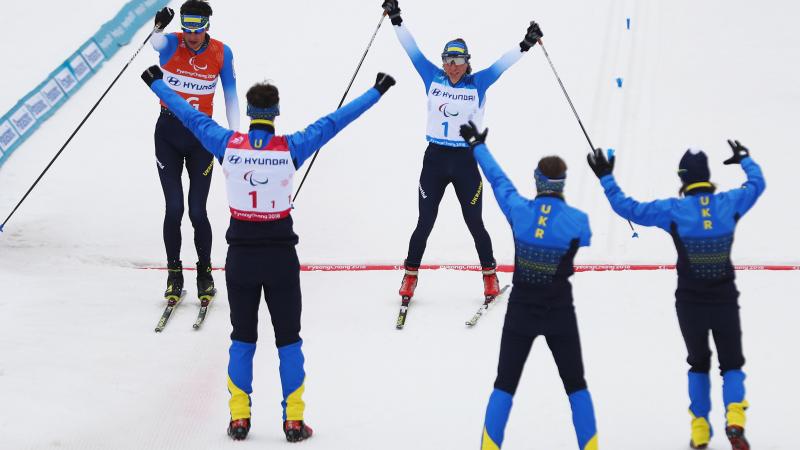 A woman crosses the finish line with raised hands and two sticks in them as the team cheers her on.