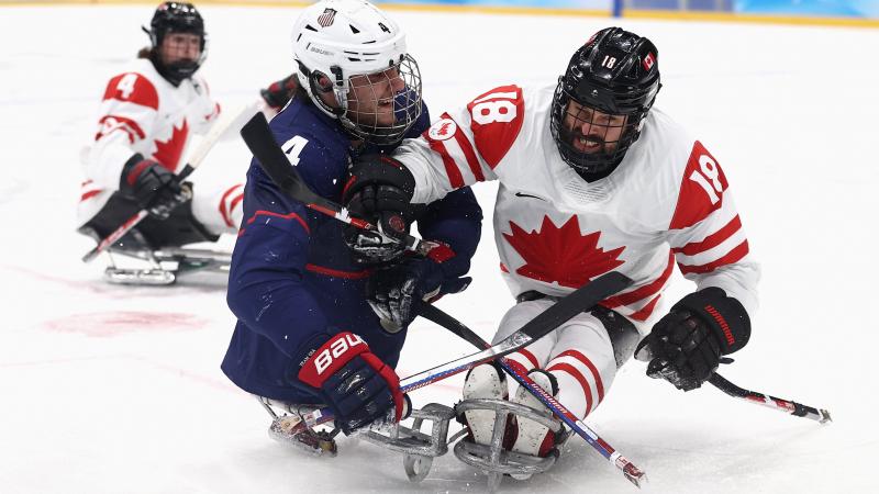 Two Para ice hockey players in full gear, one in white and the other in the blue jersey, pushing each other to gain possession of the puck.