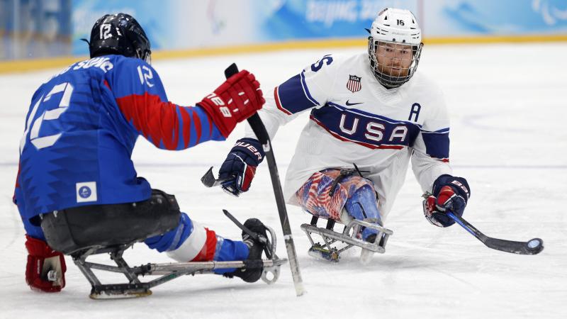 A Para ice hockey player in white and blue jersey hits the puck with a stick in left hand.