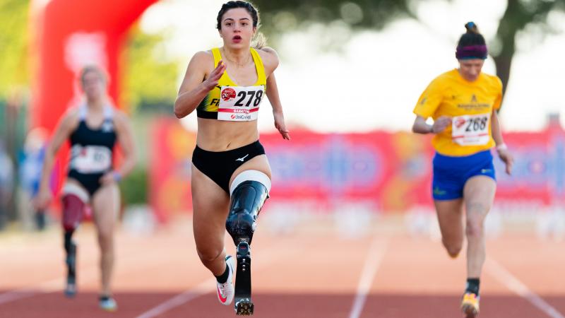 A female athlete with her left leg amputated above the knee running on the track in front of two other female athletes.