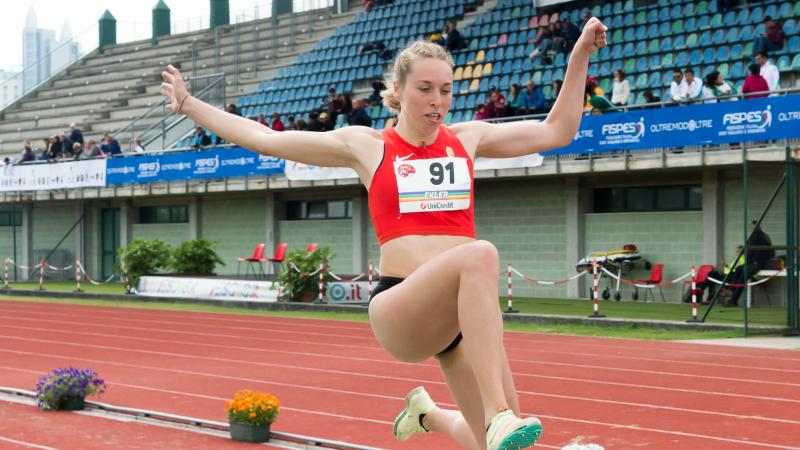 A woman in a red top jumping at the stadium with both of her hands in the air.