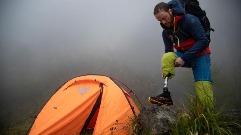 Andrea Lanfri steps on a rock as he adjusts his right prosthetic leg, with a bright orange tent on his right and heavy fog surrounding him.