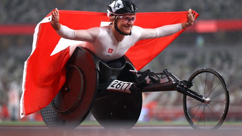 A woman in a racing wheelchair holding the Swiss flag with a smile on her face.
