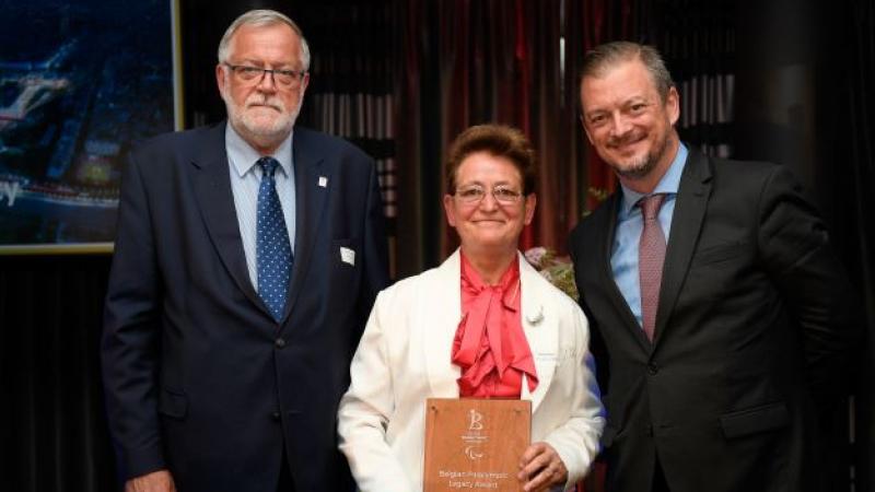 Two men in suits stand on both sides of a woman holding an awards plaque.