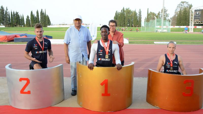 Three men on a podium with a man and a woman in the background on an athletics track
