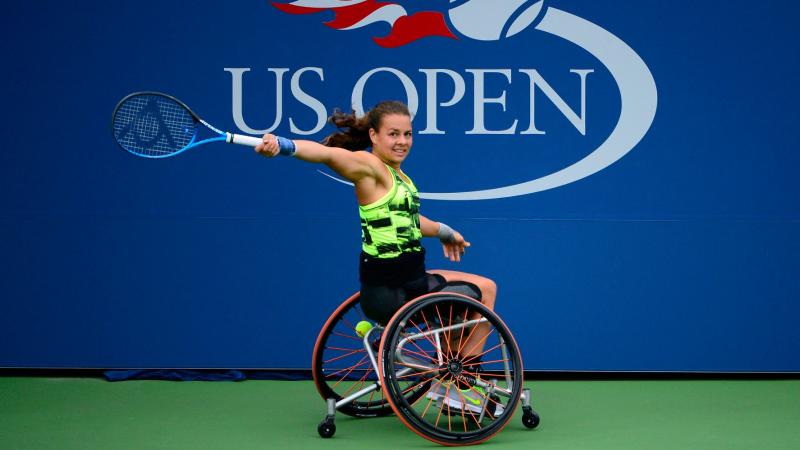 A female wheelchair player returns a shot with the words US Open written prominently on an official backdrop behind her.