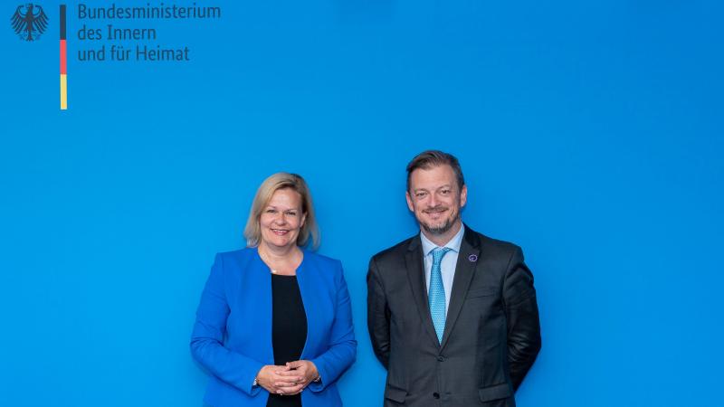 A woman and a man in business attire stand in front of an official blue backdrop.