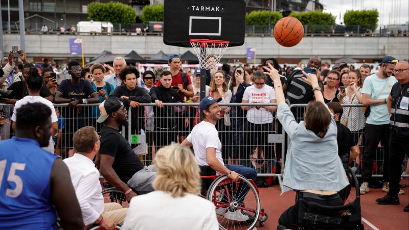 A woman sits in a wheelchair as she makes a basketball shots while a crowd of spectators looks on.