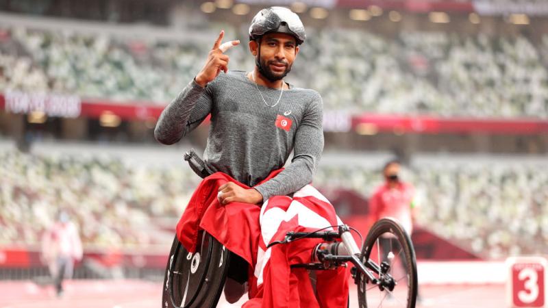 A wheelchair racer, his race chair draped in a Tunisian flag, lifts his hand in celebration on the track.