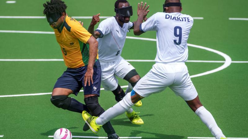 A Brazilian player breaks through a tackle from two French players in a blind football match at Tokyo 2020.