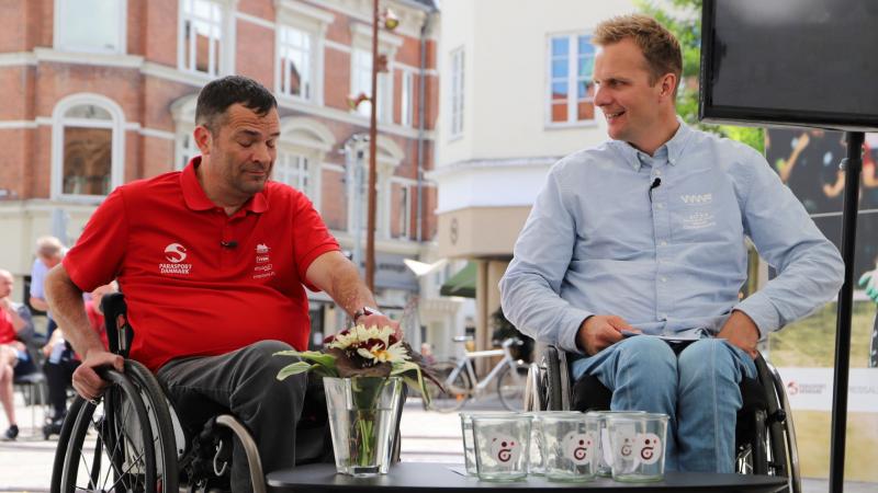 Two men in wheelchairs face a table set with glasses for the 2022 Wheelchair Rugby World Championship draw.