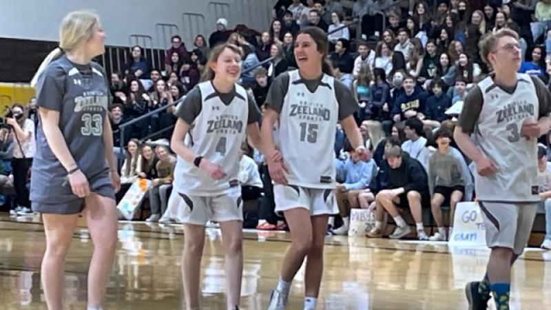 A friend guides a blind girl, both in basketball uniforms, across a basketball court in a packed stadium.