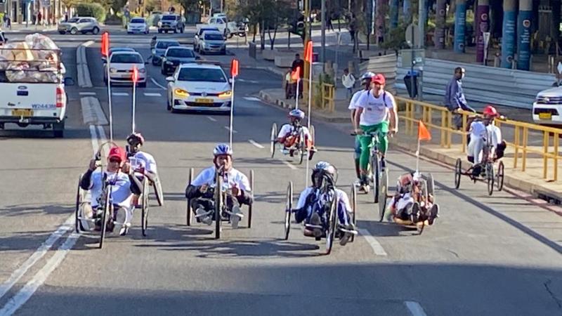 Six handcyclists ride through the city centre in Windhoek with cars following behind.