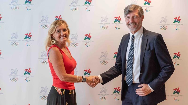 A woman and a man shake hands in front of a Milan Cortina 2026 media backdrop