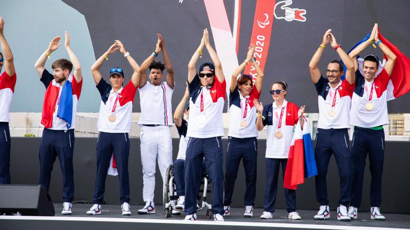 Ten people in French uniforms clap onstage with their hands raised high.