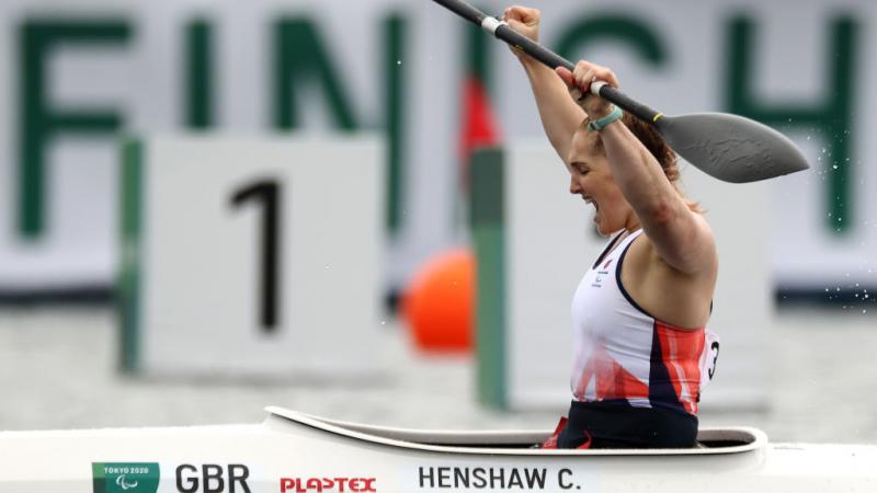 A female Para canoeist raises her paddle and yells out in celebration after crossing the finish line.