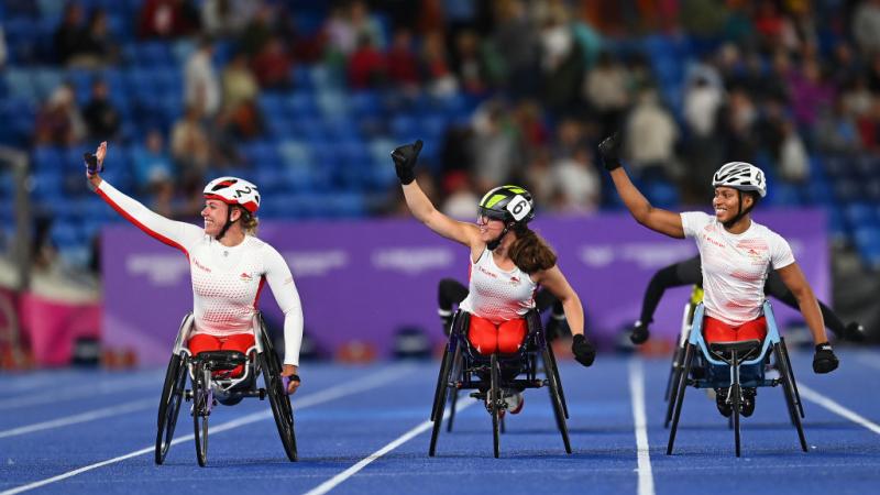 Three female wheelchair racers on a blue track in a crowded stadium
