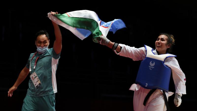 A smiling male coach and female athlete wave an Uzbek flag between them after a competition bout.