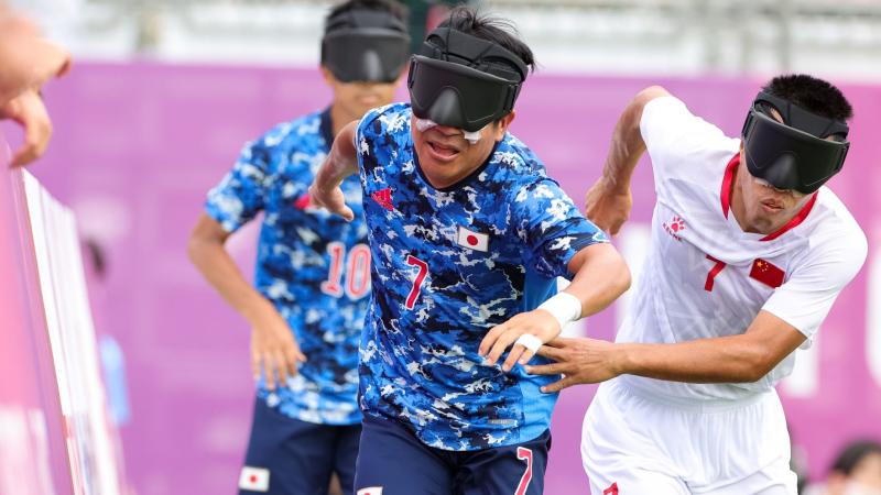 A blind football player in a blue jersey competes for the ball against a player in a white jersey at Tokyo 2020 Paralympic Games.