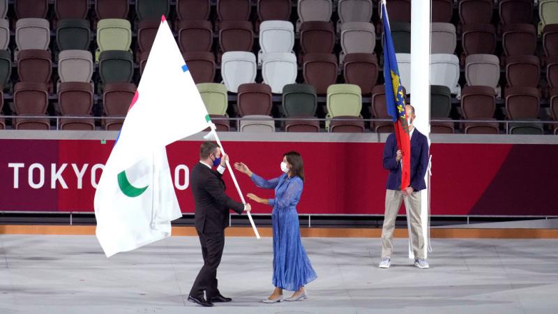 A man passes the Paralympic flag to a woman who stretches out her hands to receive it.