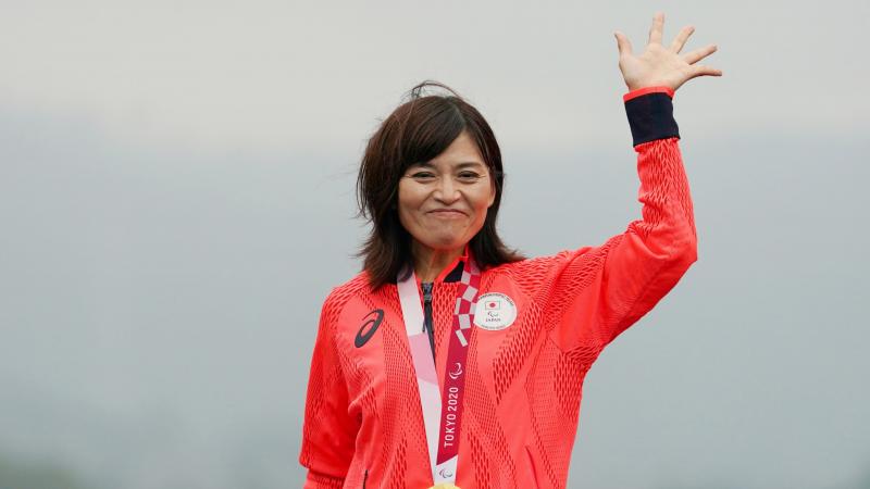 A Japanese female athlete with a gold medal around her neck waves her hand.