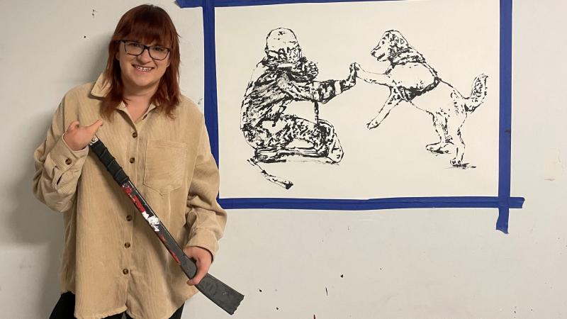 A female artist holding a Para ice hockey stick and stands in front of a painting of a female Para ice hockey player and her dog.