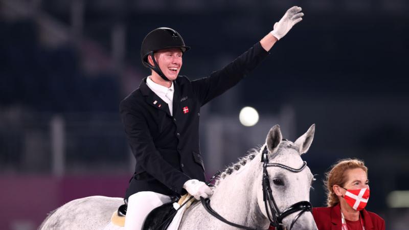 A male athlete riding a horse raises his left hand. He is accompanied by a woman wearing a red and white face mask.