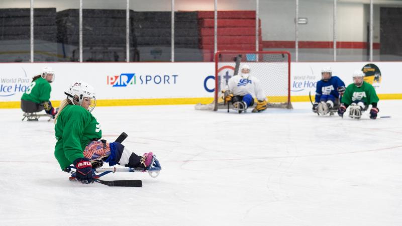 A woman in green jersey and on a sled is preparing herself to shoot the puck.