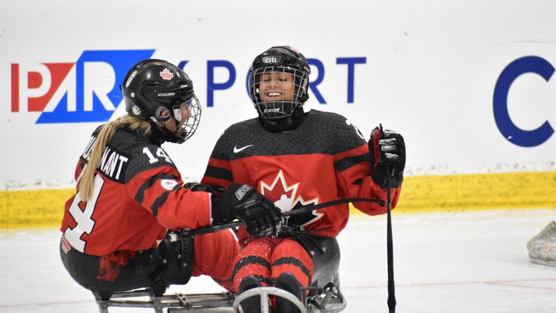 Two female Para ice hockey players on a sled smiling under their helmets in red jerseys.