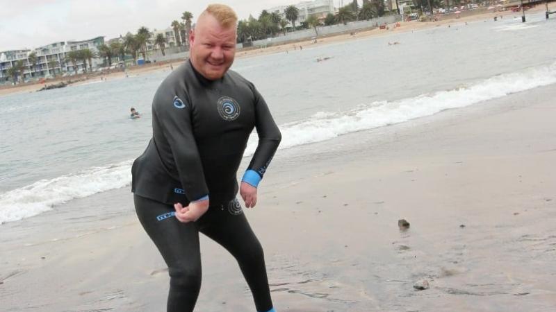 A male in a wet suit stands on the ocean shore, his feet in the water.