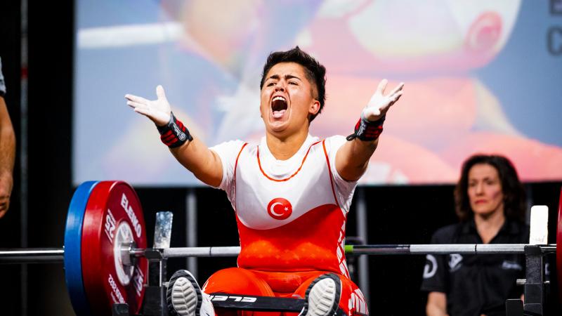 A female powerlifter raises her hands and yells in celebration while seated on a bench.