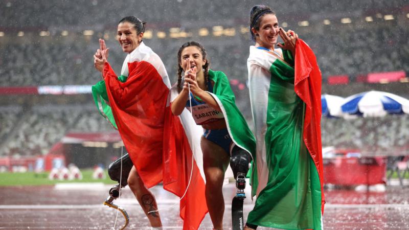 Three female athletes pose by raising two fingers in front of their faces at the Tokyo 2020 Paralympic Games.