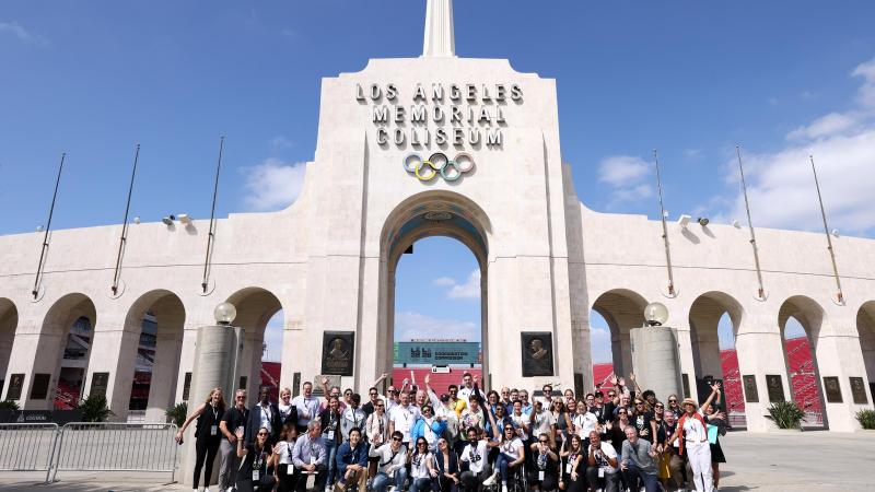 A group of people, many wearing LA28 shirts, pose for a photo in front of the Los Angeles Memorial Coliseum.