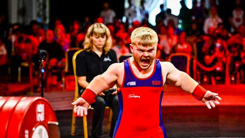 A male Para powerlifter yells in front of the barbell with his arms wide.