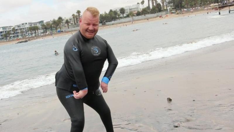 A male in a wet suit stands on the ocean shore, his feet in the water.