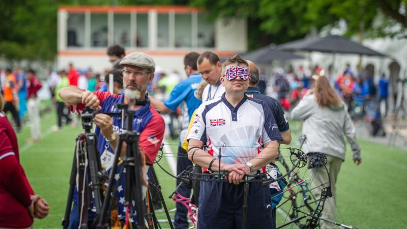 An archer in a blindfold decorated with the Union Jack rests his hands on a downturned bow.